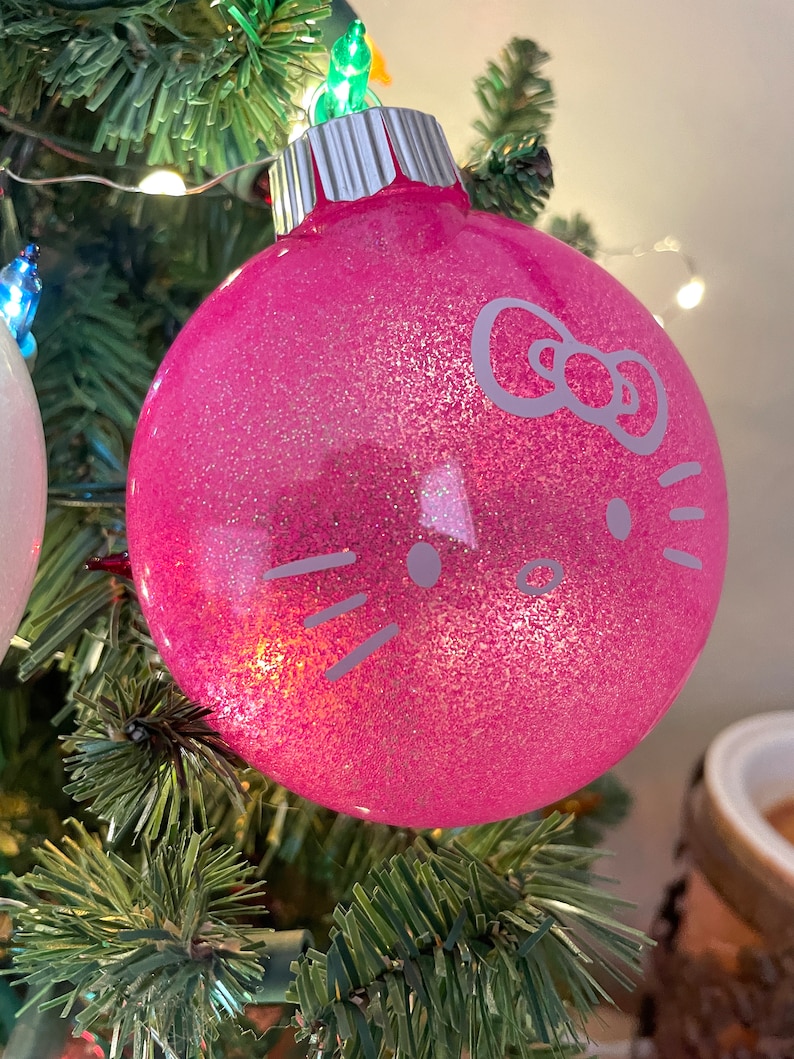 Op de afbeelding: Een roze glitter kerstbal met een zilveren dop. De ornament heeft een witte afbeelding van een kattengezicht met een strik. De bal hangt aan een groene kerstboom.
