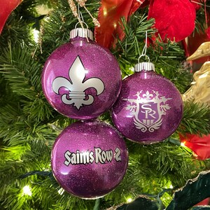 May include: Three purple glitter Christmas ornaments with silver designs and text. One ornament features a fleur-de-lis, another has a crest with "SR", and the third says "Saints Row 2". The ornaments hang on a Christmas tree.