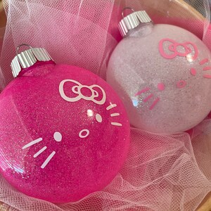 Op de afbeelding: Twee met glitter bedekte kerstballen. E&eacute;n is felroze, de andere is wit. Beide hebben een cartoon kattengezicht met een strik en snorharen. De ballen liggen in een met roze gaas gevoerde mand.