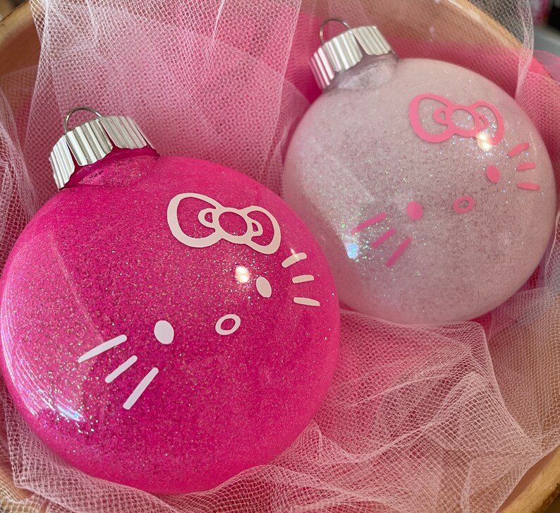 Op de afbeelding: Twee met glitter bedekte kerstballen. E&eacute;n is felroze, de andere is wit. Beide hebben een cartoon kattengezicht met een strik en snorharen. De ballen liggen in een met roze gaas gevoerde mand.