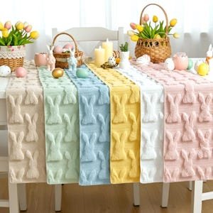 Può includere: Un'apparecchiatura da tavola con sei runner dai colori pastello, ciascuno con un motivo a coniglietto ripetuto. I runner sono beige, verde chiaro, azzurro, giallo, bianco e rosa. Decorazioni pasquali, tra cui cesti di fiori e uova decorative, sono sul tavolo.