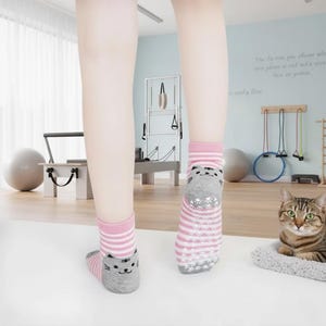 Pode incluir: Meias listradas rosa e brancas com um desenho de rosto de gato cinza. As meias têm uma aderência antiderrapante nas solas. Um gato malhado está descansando em uma cama cinza para animais de estimação. Meias de tornozelo.