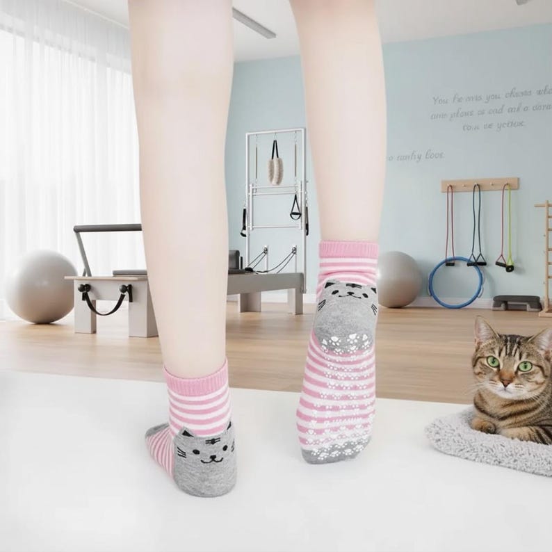 Peut inclure: Chaussettes ray&eacute;es roses et blanches avec un motif de visage de chat gris. Les chaussettes ont une adh&eacute;rence antid&eacute;rapante sur les semelles. Un chat tigr&eacute; se repose sur un lit gris pour animaux de compagnie. Chaussettes courtes.