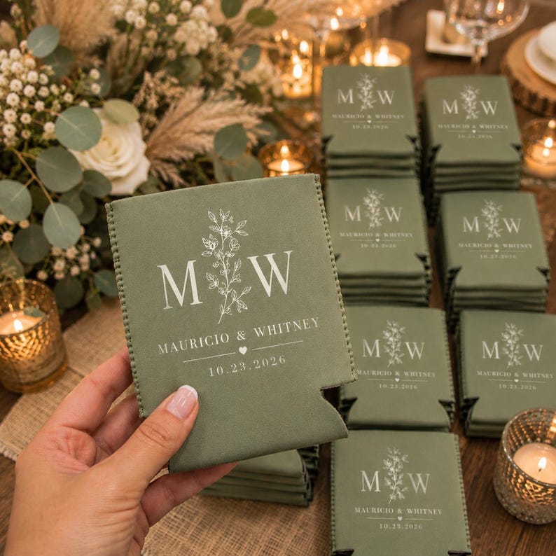 May include: A sage green foam can cooler featuring a white printed design with the letters M and W, a floral illustration, the names Mauricio & Whitney, a small heart icon, and the date 10.23.2026. Multiple identical can coolers are stacked nearby.