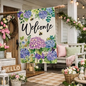 May include: A decorative outdoor flag with the word "Welcome" in black script, featuring a floral design of purple and blue hydrangeas and bees. The flag is displayed on a wooden porch with other spring decorations.