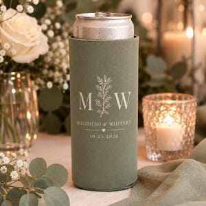 May include: A sage green slim can cooler featuring a white printed design with the letters M and W, a botanical sprig, the names Mauricio & Whitney, a small heart icon, and the date 10.23.2026, holding a silver aluminum beverage can.