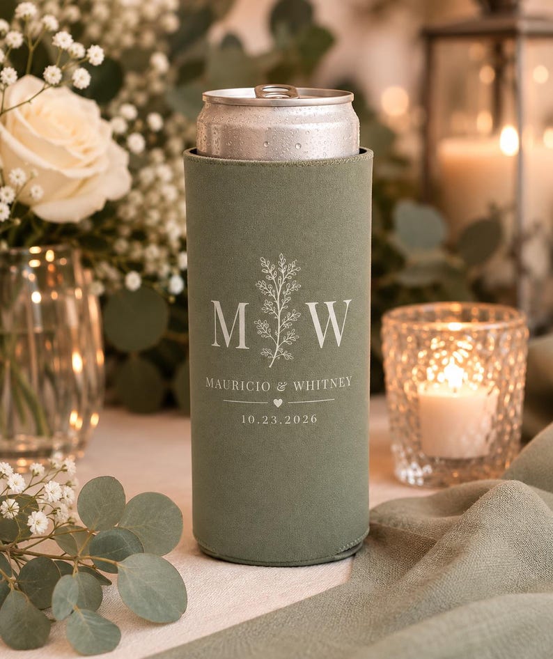 May include: A sage green slim can cooler featuring a white printed design with the letters M and W, a botanical sprig, the names Mauricio & Whitney, a small heart icon, and the date 10.23.2026, holding a silver aluminum beverage can.