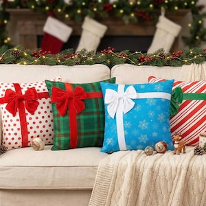 Könnte beinhalten: Vier dekorative Weihnachtskissen auf einem cremefarbenen Sofa. Die Kissen zeigen Geschenkbox-Designs mit roten Schleifen, grünem Karomuster, weißen Schleifen, blauen Schneeflocken und rot-weißen Streifen. Eine festliche Urlaubsszene.