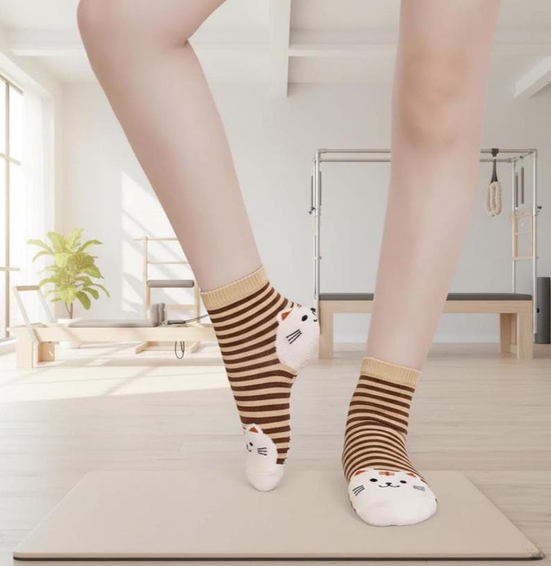 Peut inclure: Une paire de chaussettes avec un motif de visage de chat. Les chaussettes ont des rayures horizontales marron et beige et un visage de chat blanc sur l'orteil. Les chaussettes sont port&eacute;es sur une surface de couleur claire.