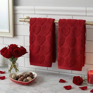 Pode incluir: Duas toalhas de banho vermelhas com padrões de coração penduradas numa barra dourada. Um buquê de rosas vermelhas, um prato em forma de coração com chocolates e uma vela acesa estão sobre a bancada. A cena evoca um tema romântico de Dia dos Namorados.