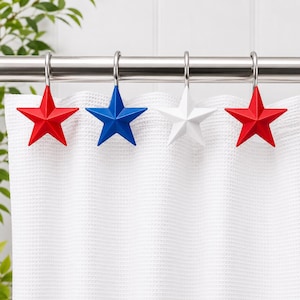 May include: A set of four star-shaped shower curtain hooks in red, white, and blue. The hooks are made of a shiny metal and hang from a silver rod. The shower curtain is white and has a waffle weave texture.