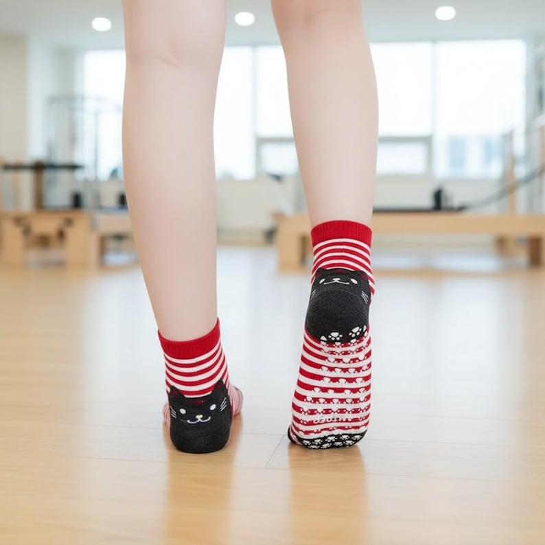 Peut inclure: Chaussettes ray&eacute;es rouges et blanches avec un motif de visage de chat noir sur le talon. Les chaussettes ont un bord rouge et une adh&eacute;rence antid&eacute;rapante sur la semelle. Les chaussettes sont sur un sol en bois.