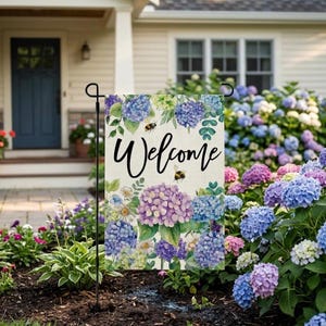 Puede incluir: Una bandera de jardín con la palabra "Welcome" en escritura negra, rodeada de flores de hortensia azules, moradas y blancas y dos abejorros. La bandera se muestra en un soporte de metal negro en un jardín.
