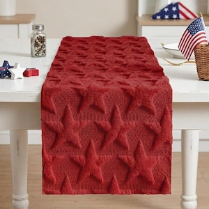 May include: A red table runner with a star pattern. The runner is placed on a white table, with decorative stars and American flags. The table runner is made of a soft, textured material.