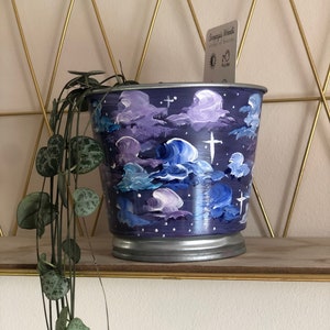 May include: A painted metal planter with a night sky design featuring clouds, stars, and crosses. The planter is filled with a trailing plant with green leaves.