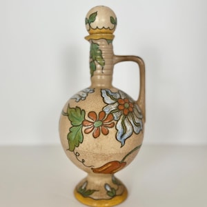 May include: A tan ceramic bottle with a round body and a handle, decorated with colourful floral designs in orange, blue, and green. The bottle has a matching stopper and sits on a pedestal base with a yellow trim. The design is vintage.