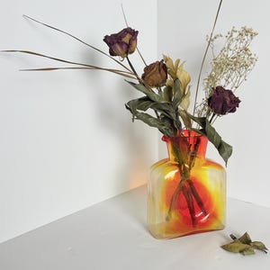 Puede incluir: Un jarrón cuadrado de vidrio con un degradado de amarillo, naranja y rojo. El jarrón contiene flores y tallos secos, incluyendo rosas moradas oscuras y delicadas flores blancas. El jarrón está colocado en una esquina.