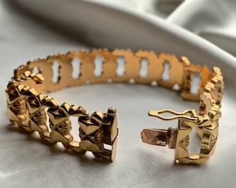May include: A gold bracelet with a detailed, intricate design. The bracelet is open and the clasp is visible.