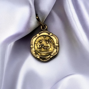 May include: A gold-colored pendant with a detailed relief of figures within a floral-edged frame. The pendant hangs from a small loop and is set against a backdrop of white, flowing fabric. The pendant's design is intricate and ornate.