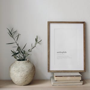 May include: A framed print with the word "anthophile" and its definition, displayed on a wooden shelf with a stack of books. A vase with a textured surface holds a sprig of green foliage.