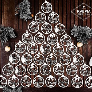 May include: A white wooden Christmas tree ornament set with 25 ornaments featuring various names in a white script font. The ornaments are arranged in a tree shape on a wooden surface. The text "KVEPIA MEDIENA" is visible in the top right corner of the image.
