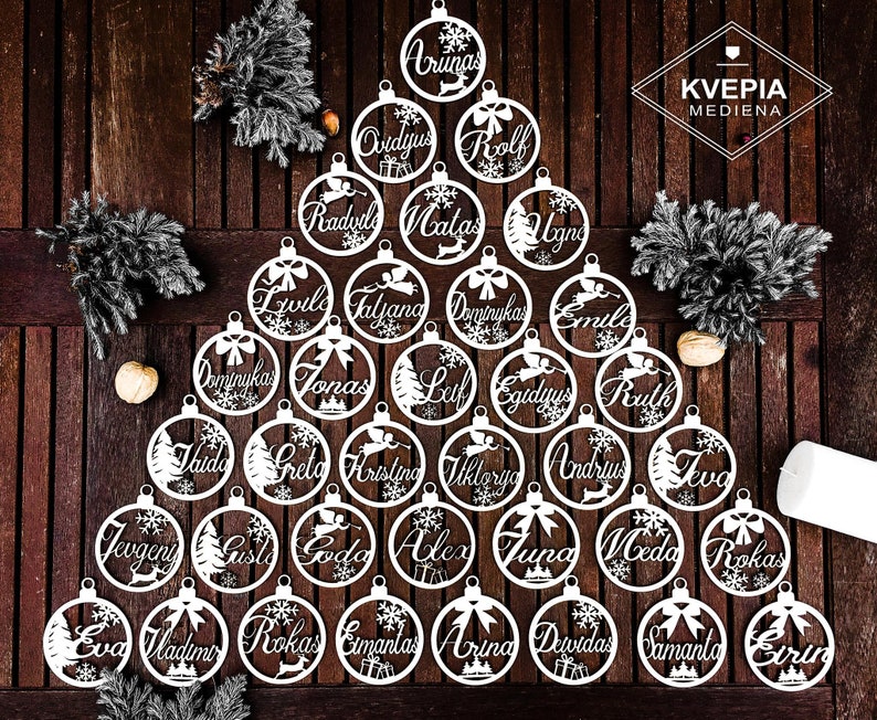 May include: A white wooden Christmas tree ornament set with 25 ornaments featuring various names in a white script font. The ornaments are arranged in a tree shape on a wooden surface. The text "KVEPIA MEDIENA" is visible in the top right corner of the image.
