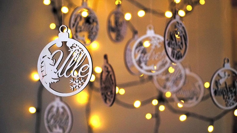 May include: White wooden Christmas ornaments with the word "Ville" and other words in a decorative font. The ornaments are hanging from a string of lights.