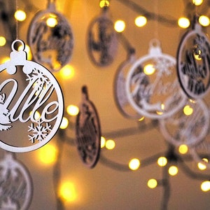 May include: White wooden Christmas ornaments with the word "Ville" and other words in a decorative font. The ornaments are hanging from a string of lights.