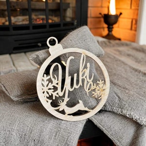 May include: A wooden Christmas ornament with a reindeer and snowflake design. The ornament is personalized with the name "Yuki".