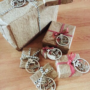 May include: A collection of wrapped gifts with personalized wooden ornaments. Each ornament features a different name, including "Vaida", "Neilas", "Agne", "Karolina", "Andrius", and "Santa". The ornaments are tied with red and white ribbon and brown and white polka dot ribbon.