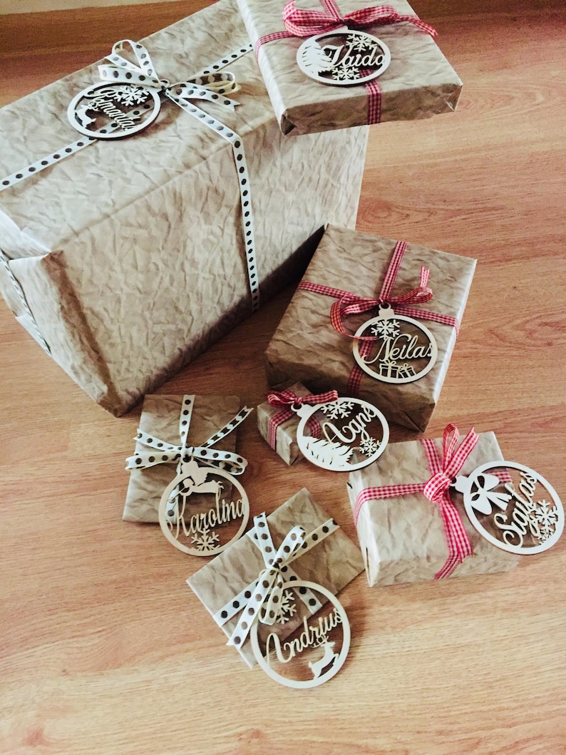 May include: A collection of wrapped gifts with personalized wooden ornaments. Each ornament features a different name, including "Vaida", "Neilas", "Agne", "Karolina", "Andrius", and "Santa". The ornaments are tied with red and white ribbon and brown and white polka dot ribbon.