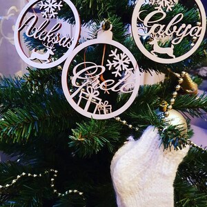 May include: Three wooden Christmas ornaments with names "Aleksandra", "Gabriela", and "Greta" written on them. The ornaments are hanging on a green Christmas tree with white lights and a white knitted stocking.