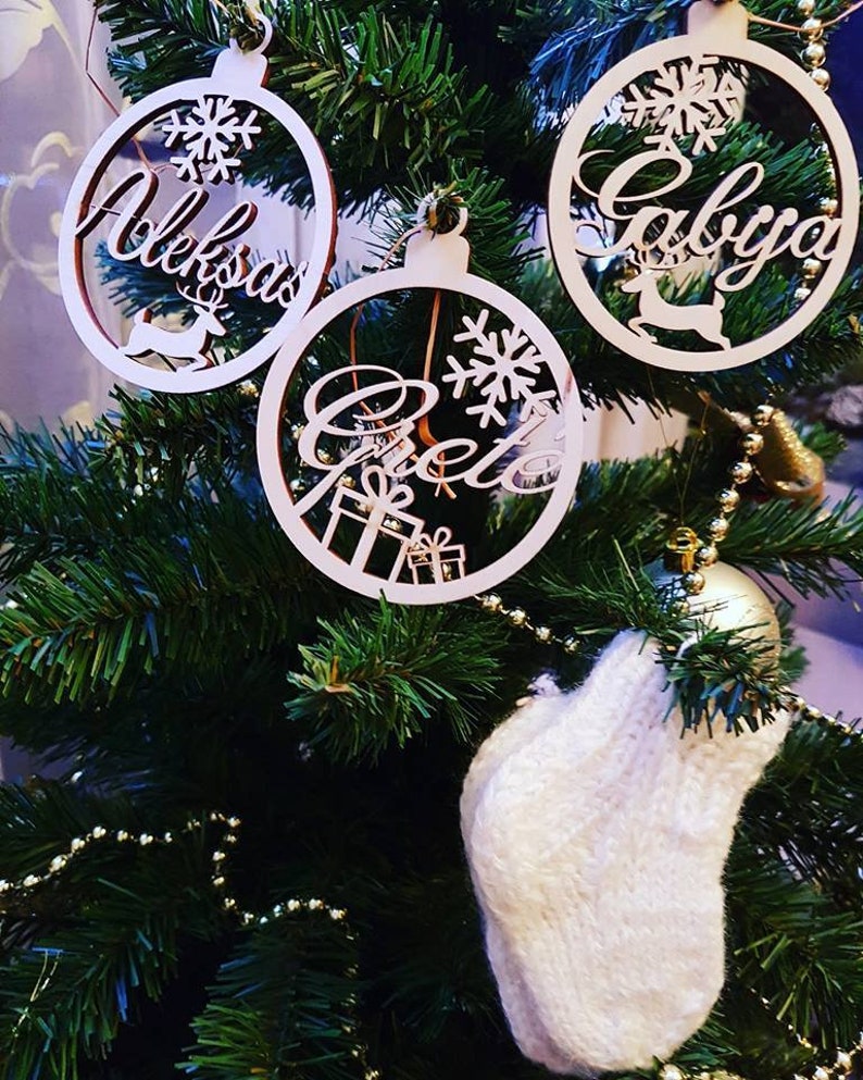 May include: Three wooden Christmas ornaments with names "Aleksandra", "Gabriela", and "Greta" written on them. The ornaments are hanging on a green Christmas tree with white lights and a white knitted stocking.