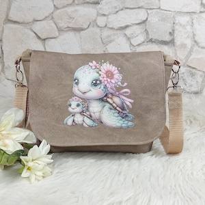 Könnte beinhalten: Eine braune Canvas-Messenger-Tasche mit einem bedruckten Design aus zwei Comic-Schildkröten. Die größere Schildkröte trägt eine rosafarbene Blumenkrone und die kleinere Schildkröte sitzt auf ihrem Rücken. Die Tasche hat einen verstellbaren braunen Riemen.