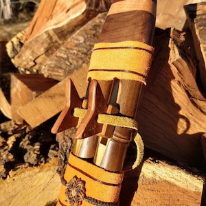 May include: A handcrafted wooden flute with a multi-toned wood body and a leather wrap. The flute features a decorative star-shaped element and is accented with yellow leather and dark brown accents. The instrument is set against a backdrop of stacked firewood.