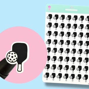 May include: Sheet of stickers featuring a black pickleball paddle and ball design. The stickers are on a white background with a light blue and pink background. The stickers are labeled with the text "jelly studio designs".