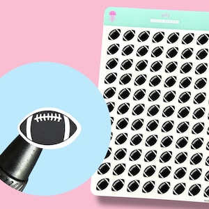May include: Sheet of stickers featuring black and white footballs. A single sticker is held up by a black object. The background is pink and light blue. The stickers are for crafts and sports-themed projects.