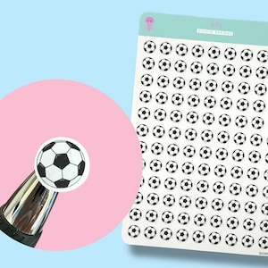 May include: A sheet of soccer ball stickers with a close-up of a single sticker on a pen. The stickers are black and white, and the sheet has a light green border with the text "jelly STUDIO DESIGNS". The background is light blue and pink.