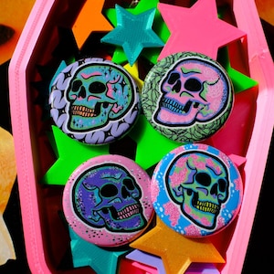 May include: Four round skull-themed buttons in a pink coffin-shaped container with star-shaped decorations. The buttons feature colorful skull designs with floral and geometric patterns in shades of pink, blue, green, and purple.