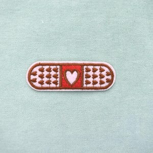 Peut inclure: Écusson brodé en forme de pansement, avec un cœur blanc au centre. Le pansement a une bordure marron et est décoré de détails marron et roses. Le fond est un tissu vert clair.