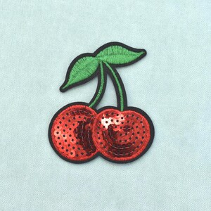 May include: A cherry-shaped iron-on patch featuring two bright red sequined fruits with green embroidered leaves and stems. The entire design is outlined in a thick black border, creating a distinct shape that stands out against the light background.