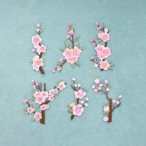 Peut inclure: Un ensemble de six écussons brodés représentant des branches de cerisier en fleurs avec des pétales roses, des feuilles vertes et des tiges brunes. Chaque écusson présente une disposition unique de fleurs et de bourgeons détaillés.