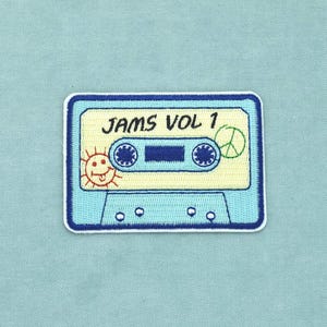 Peut inclure: Un écusson brodé rectangulaire en forme de cassette audio avec une bordure bleu clair. Le centre présente le texte JAMS VOL 1 en noir, une petite icône de soleil orange à gauche et un signe de paix vert à droite sur un fond crème.