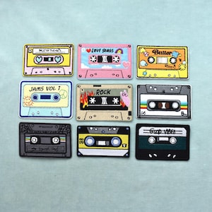 May include: Nine embroidered patches in the shape of cassette tapes. Each patch features a different design, including text like "BEST OF THE 80'S", "LOVE SONGS", "Butter", "JAMS VOL 1", "ROCK", and "GOOD VIBES". The patches are in various colors.
