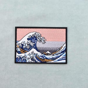 Può includere: Toppa ricamata raffigurante una grande onda oceanica con creste bianche e acqua blu, su uno sfondo rosa e grigio. La toppa ha un bordo nero ed è di forma rettangolare. Il design è ispirato all'arte giapponese.