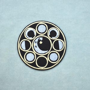 May include: Embroidered patch showing the phases of the moon. The design features a black circle with a gold border, enclosing light blue and white moon phases, and a crescent moon with stars. The patch is circular and has an iron-on backing.