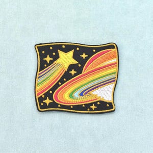 Könnte beinhalten: Ein rechteckiger bestickter Aufnäher mit schwarzem Hintergrund, der einen gelben Sternschnuppen-Kometen mit Regenbogen-Schweif und einen großen Planeten mit Regenbogen-Ringen zeigt. Das Design enthält mehrere kleine gelbe vierzackige Sterne.