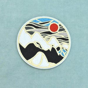 May include: A circular embroidered patch featuring a landscape design with white snow-capped mountains, black mountain bases, and a bright red sun set against a sky of blue and grey wavy lines, all enclosed within a thin white stitched border.