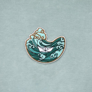 Peut inclure: Un écusson brodé représentant un bateau en papier blanc flottant sur des vagues stylisées. Les vagues sont représentées dans des tons de bleu sarcelle, vert foncé et bleu clair avec une bordure brune soulignant la forme du motif.