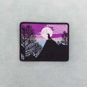 Peut inclure: Écusson brodé représentant la silhouette d'un chien sur une colline, sous une pleine lune. Le ciel est dégradé du violet au rose, avec des arbres de chaque côté. L'écusson est rectangulaire avec un bord noir.